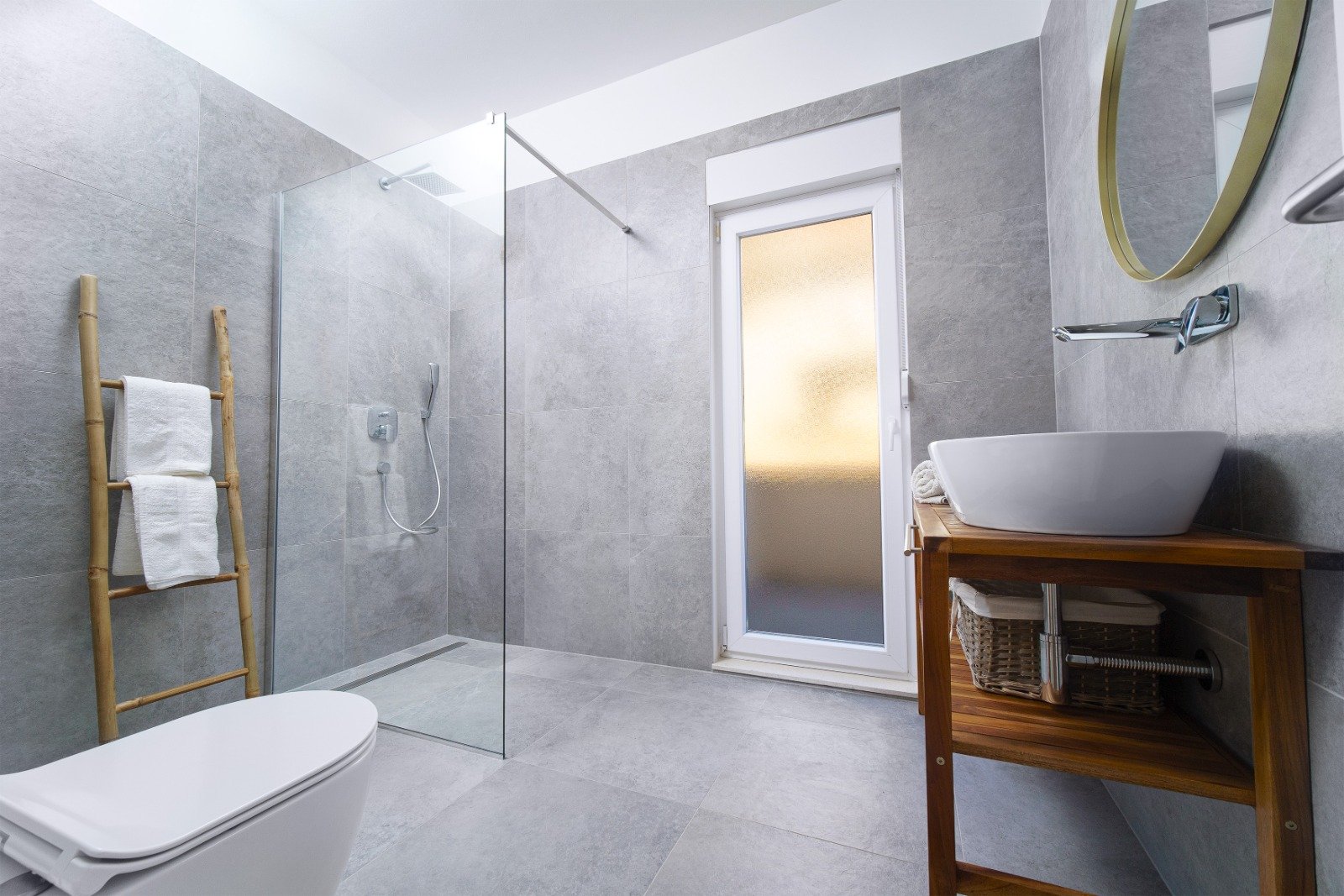 Bright grey-tiled bathroom with a glass walk-in shower, wooden basin stand and ladder towel rail.