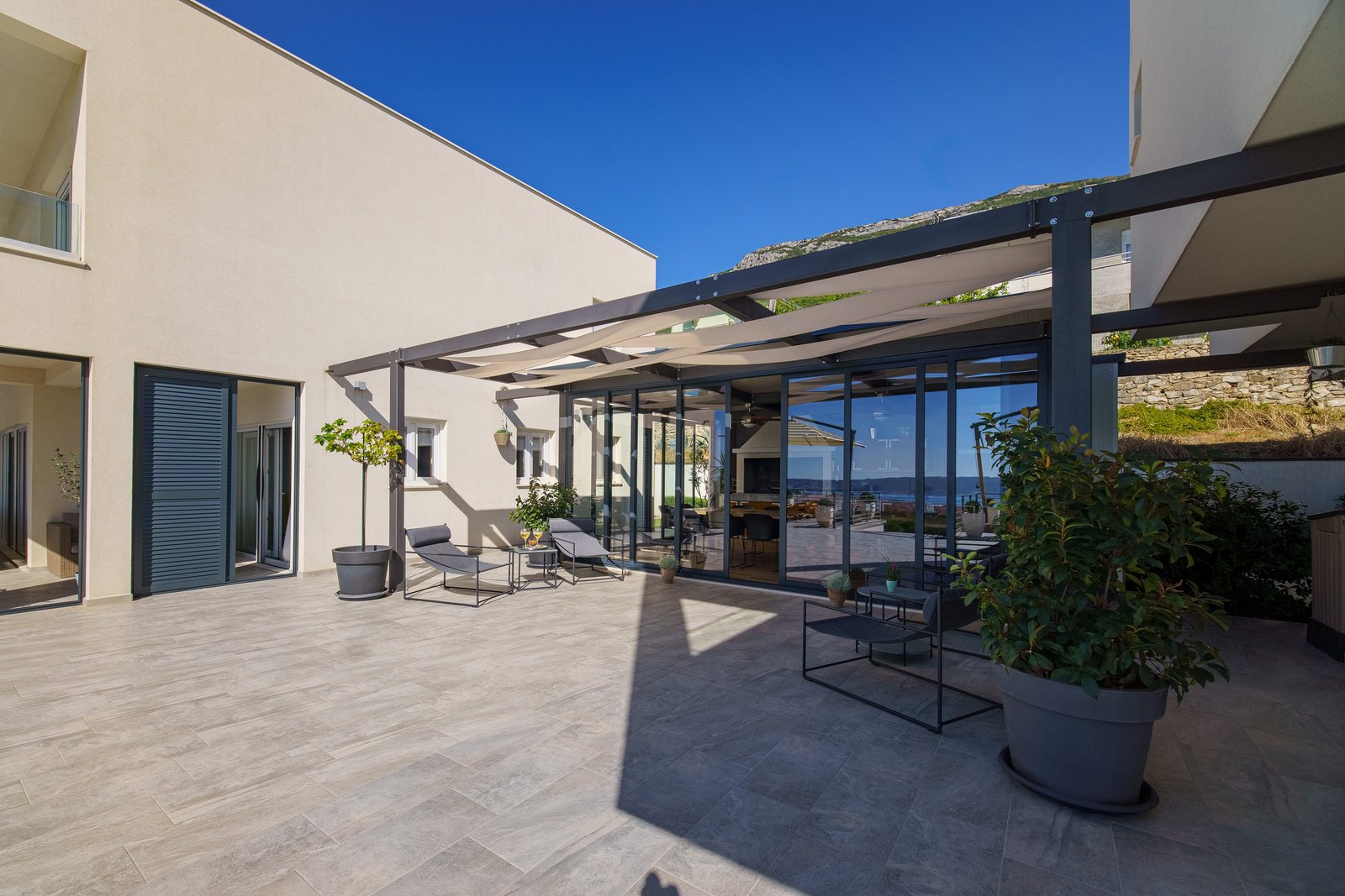 Paved courtyard between the villas with a fabric-shaded pergola, potted trees and chairs outside the glass dining pavilion.