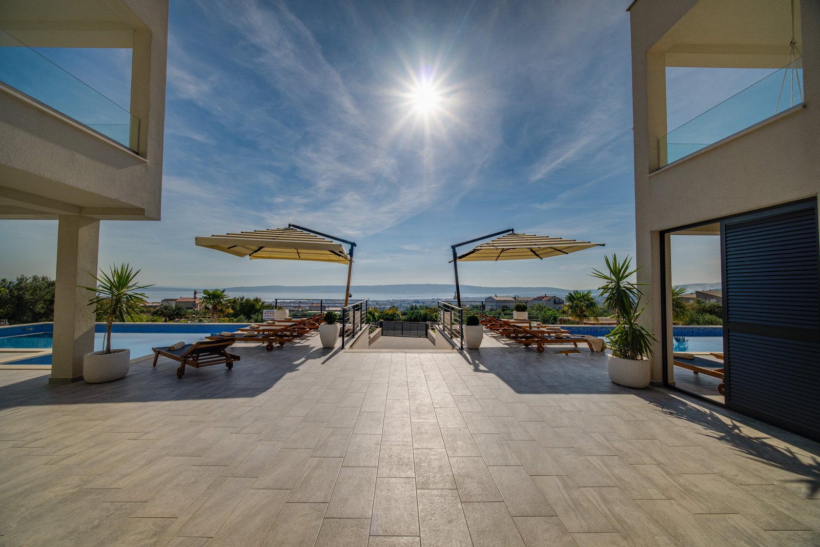 View between the two villas toward paired pool terraces with loungers and parasols, central steps and the Adriatic horizon.