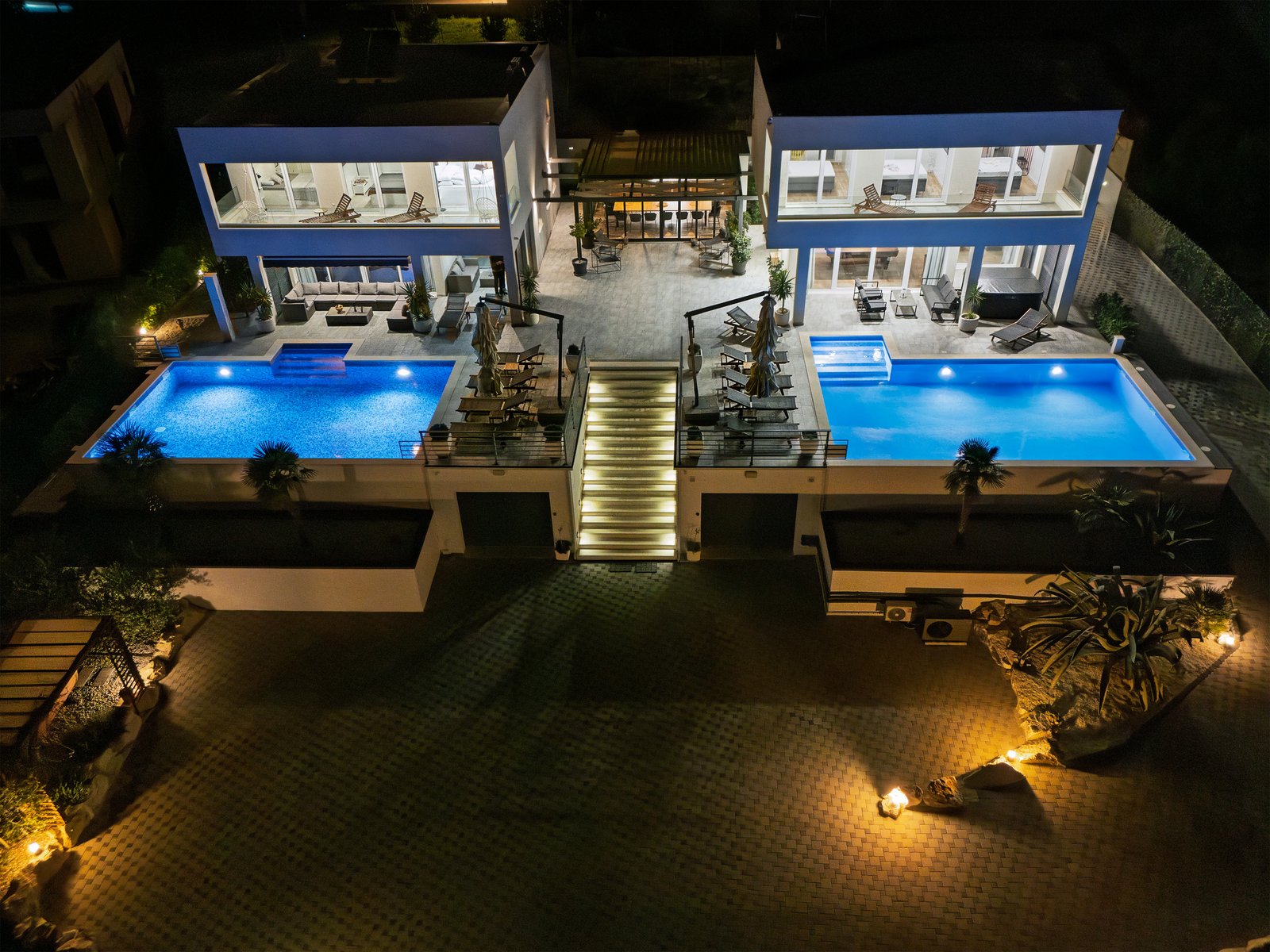 Nighttime aerial view of both villas and blue-lit pools, with illuminated central stairs, courtyard and garden borders.