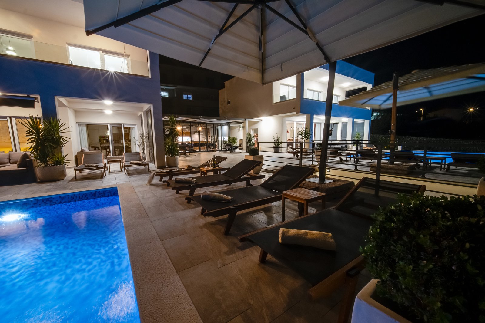 Poolside loungers beneath large parasols at night, with both villas and the central dining pavilion illuminated.