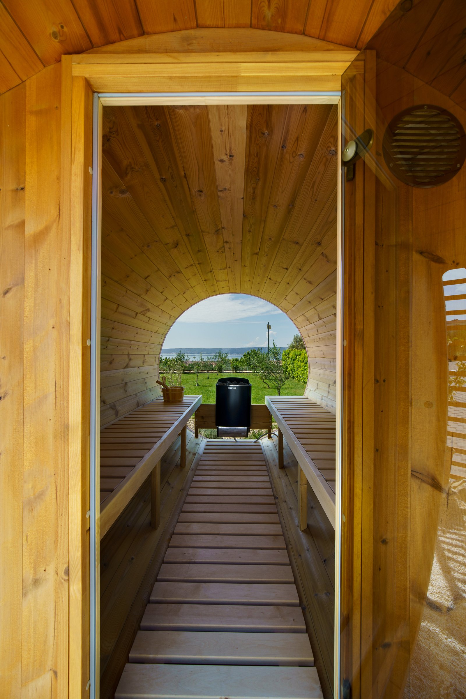 Wood-lined barrel sauna with benches and a heater, looking through a round window toward the garden and sea.