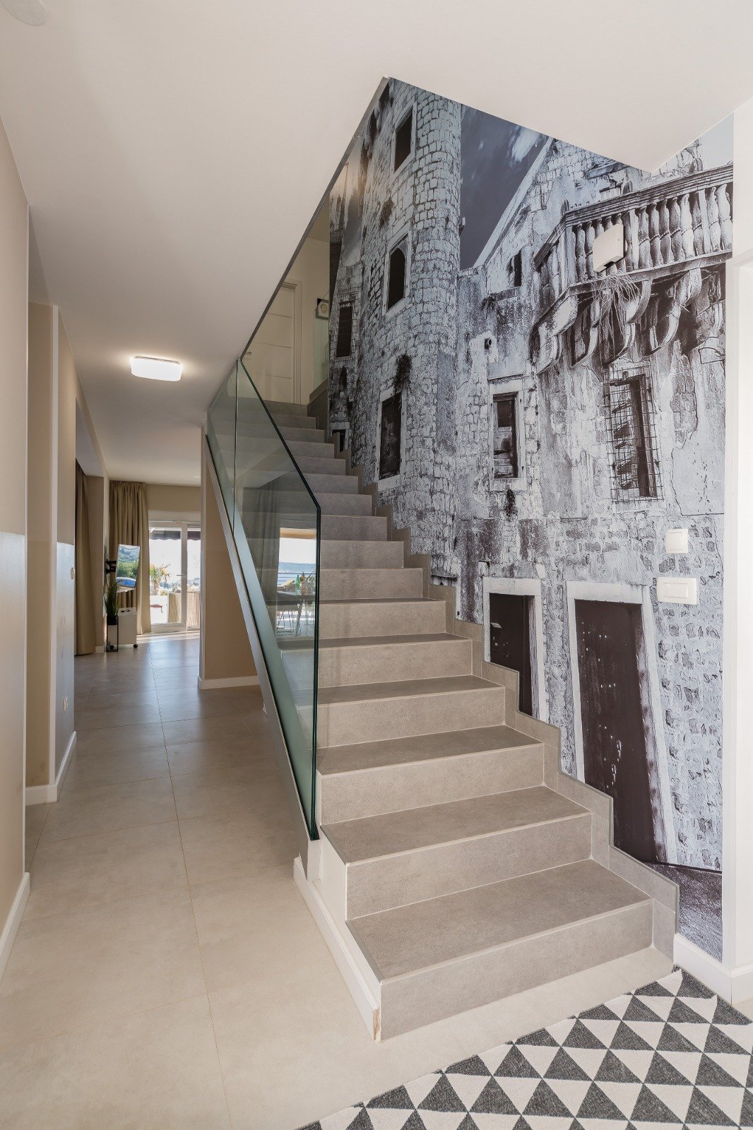 Interior staircase with a glass balustrade beside a large monochrome mural of stone architecture.